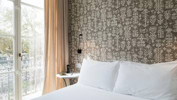Quarto de hotel com uma grande janela, cama com roupa de cama branca, mesa pequena com abajur e papel de parede estampado.