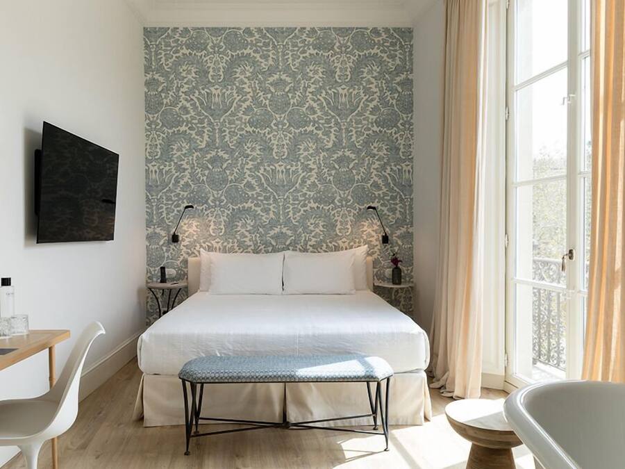 Un dormitorio con una cama grande, mesitas de noche, un escritorio, una silla, un banco, una bañera y una ventana con cortinas.