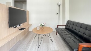 A modern living room with a flat-screen TV mounted on a wooden wall, a black leather sofa, a round wooden coffee table, and a floor lamp. - Affetto hostel (Taichung)
