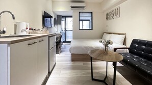 A modern hotel room with a kitchenette, a bed with white linens, a black leather sofa, and a small round table with a vase. - Affetto hostel (Taichung)
