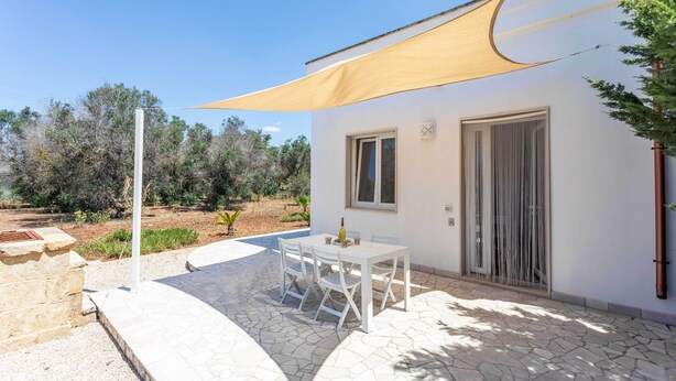 Eine Terrasse mit Tisch und Stühlen, ein weißes Gebäude mit Tür und Fenster sowie ein großes Sonnensegel.