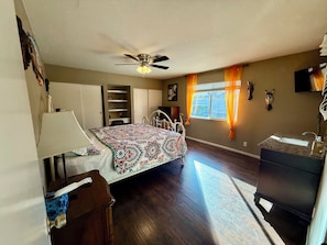 A bedroom with a bed, a desk, a wardrobe, a ceiling fan, a window with curtains, and a bathroom with a sink and mirror.