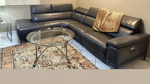 A modern living room with a black leather sectional sofa, a glass coffee table, a patterned rug, a decorative wall piece, and a painting.