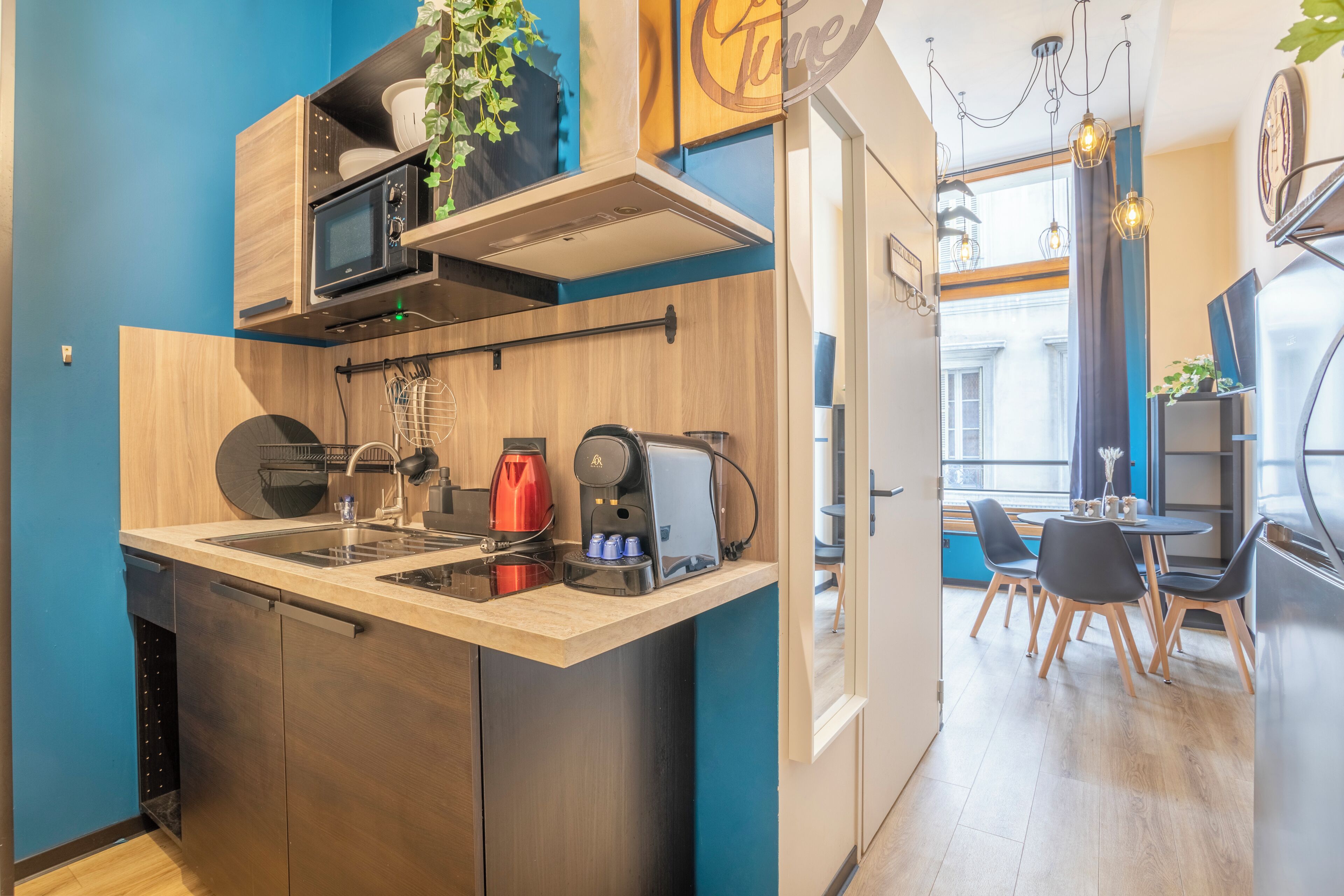 Een moderne keuken met een ingebouwde magnetron, koffiezetapparaat en spoelbak. Een eethoek met een tafel en stoelen is zichtbaar door een open deur.