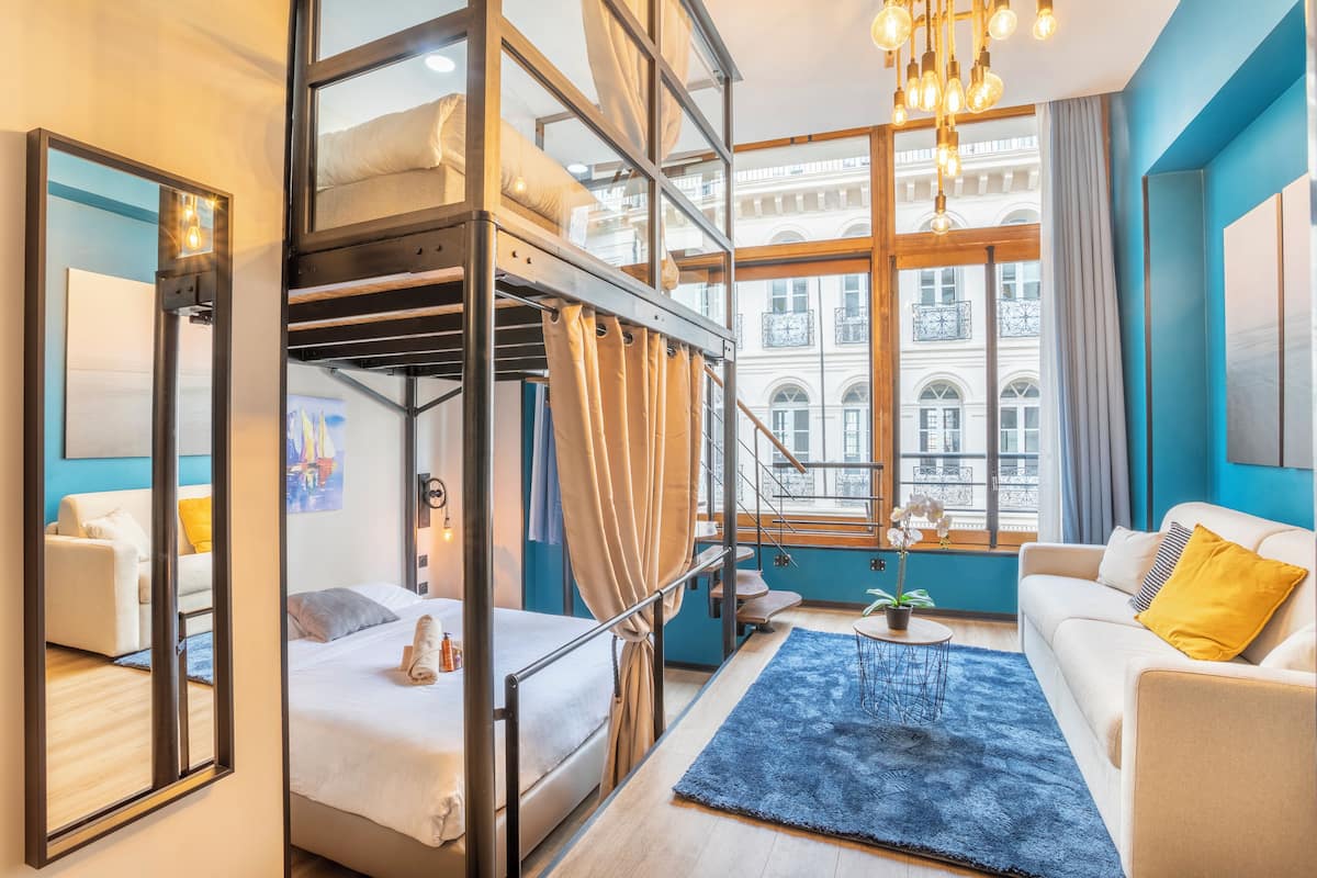 Una habitación de hotel moderna con una cama litera, un sofá, una mesa de centro y un amplio ventanal con vistas a un edificio.