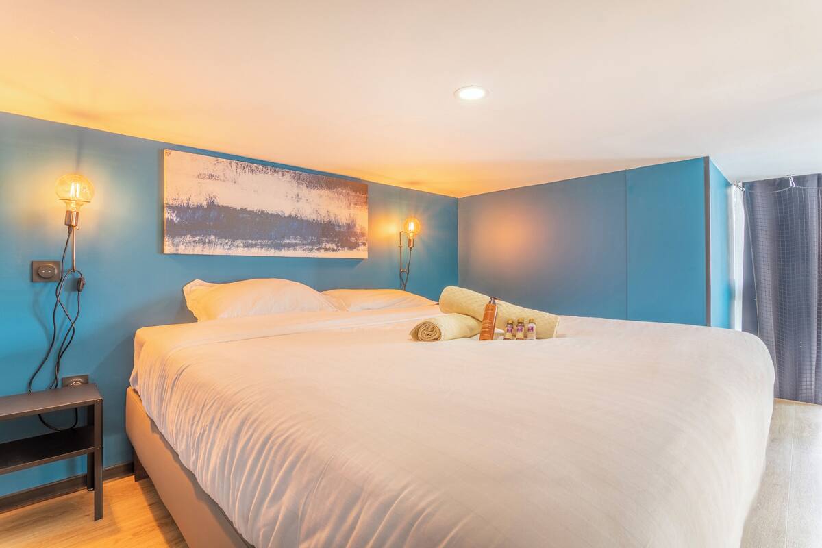 Una cama bien hecha con sábanas blancas, una mesita de noche con una lámpara y un cuadro en la pared.
