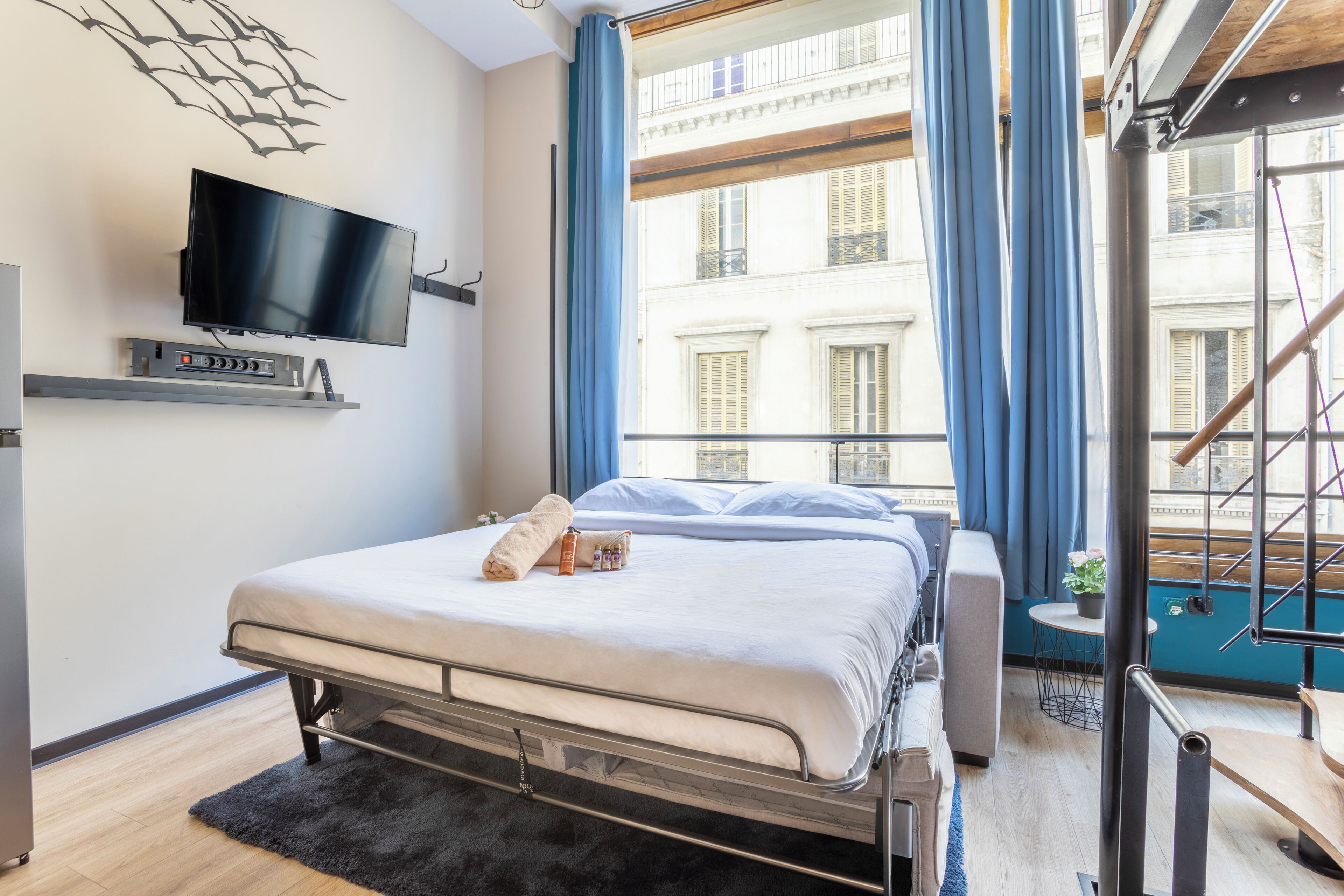Une chambre d’hôtel moderne équipée d’un lit, d’un téléviseur à écran plat, d’une petite table avec une plante et offrant une vue sur un immeuble à travers de grandes fenêtres.