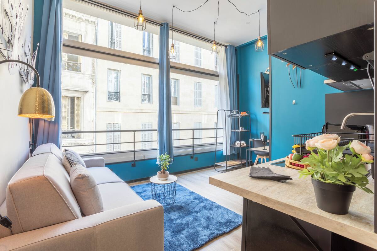 Una sala de estar moderna con un sofá, una mesa de comedor, un tapete azul y vistas de un edificio a través de amplios ventanales.