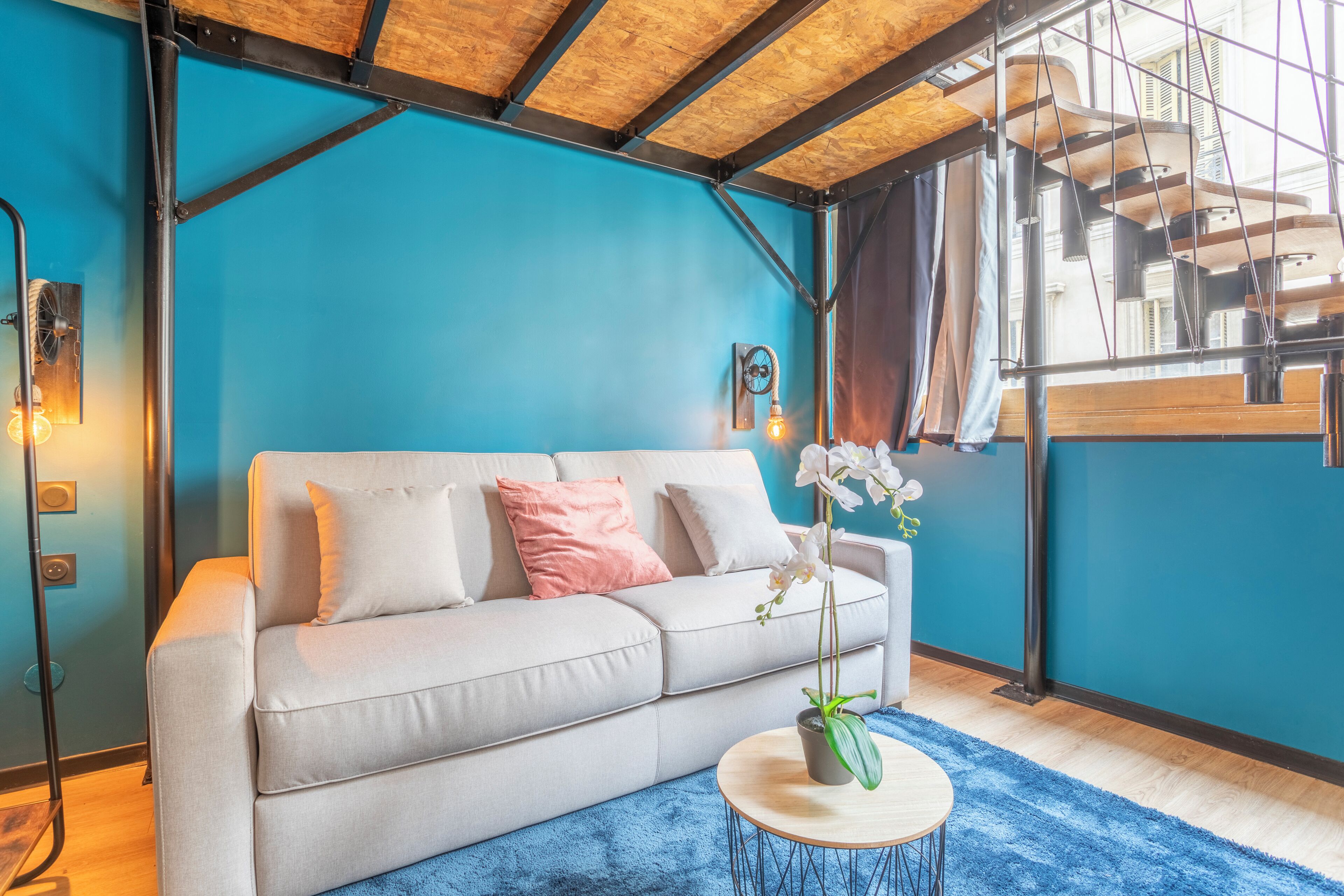 Un salon moderne avec un canapé gris clair, un tapis bleu et une orchidée en pot.