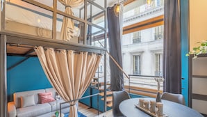 Una habitación moderna estilo loft con cama entrepiso, zona de comedor con mesa redonda y sofá con vista a la ciudad.