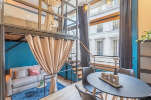 A modern loft-style room with a loft bed, a dining area with a round table, and a sofa with a view of the city.