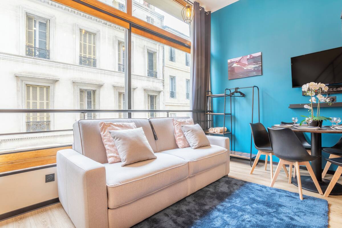 Una sala de estar moderna con sofá, mesa de comedor, sillas, televisor y vista a un edificio a través de amplias ventanas.