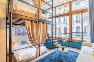 A modern hotel room with a bunk bed, a sofa, a small table with a plant, and a large window offering a view of a building with balconies.