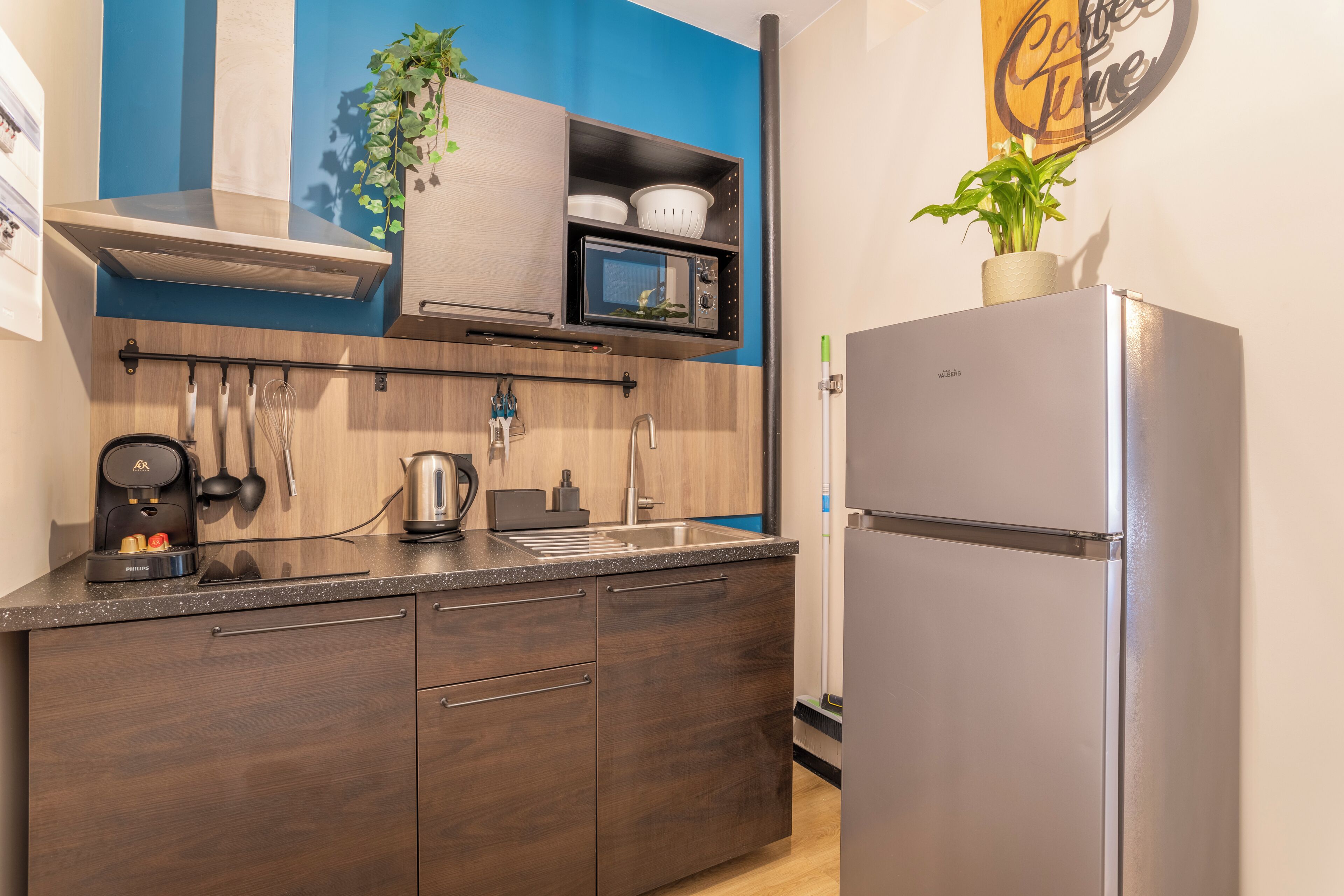 Eine moderne Küche mit einem Edelstahl-Kühlschrank, einer Mikrowelle und einer Kaffeemaschine. Es gibt einen Holzkasten mit einem Küchenutensilienhalter und einen Blumentopf auf der Arbeitsplatte.