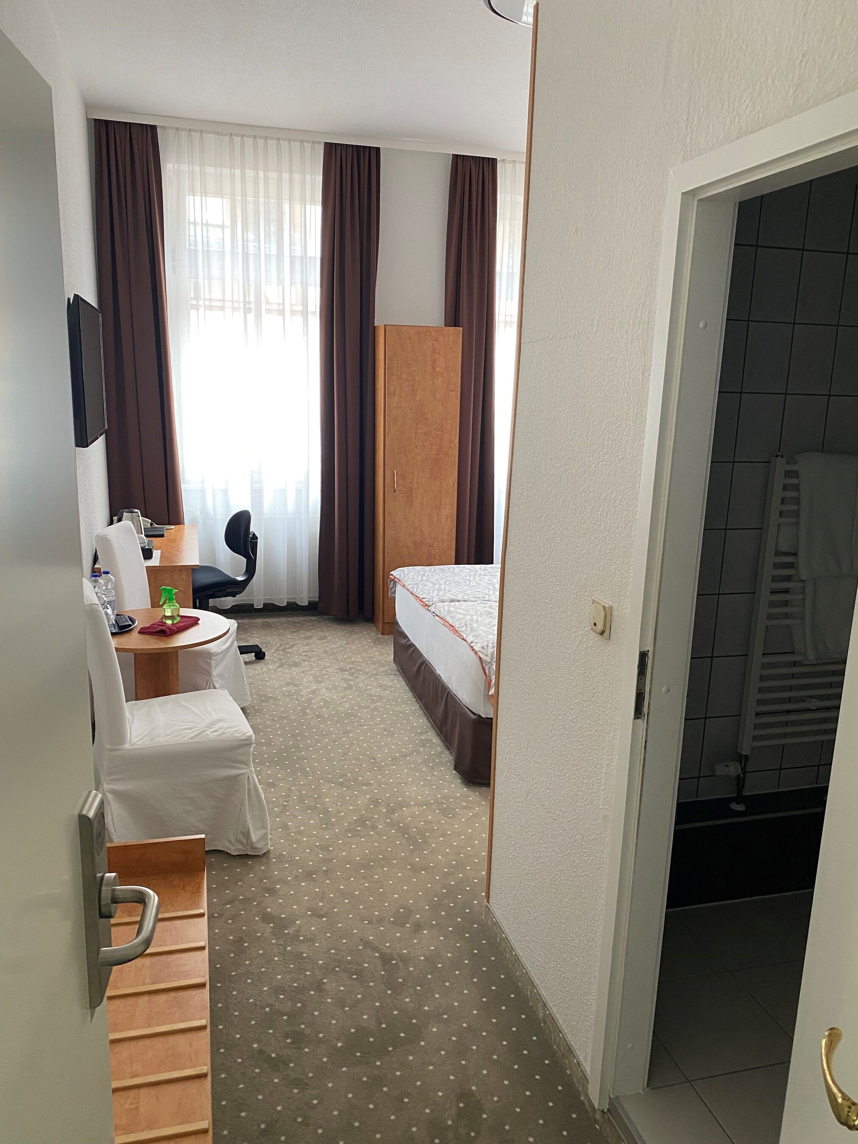 A hotel room with a bed, a desk with a chair, a wardrobe, and a bathroom visible through an open door.