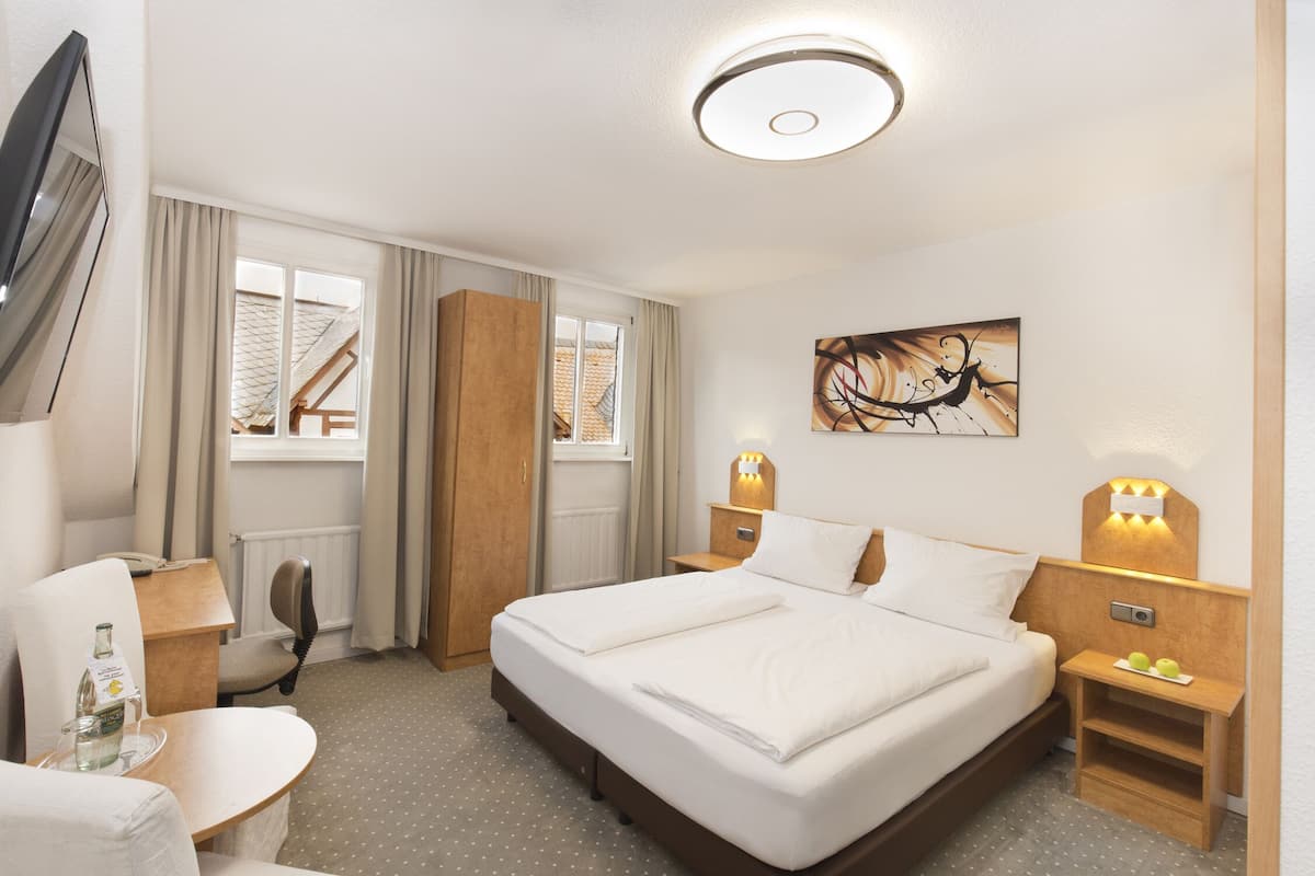 A hotel room with a bed, bedside tables, a chair, a desk, a TV, and a window with curtains.