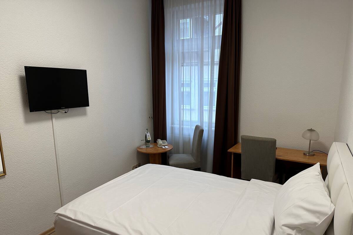 A hotel room with a bed, a TV mounted on the wall, a desk with a chair, a lamp, and a window with curtains.