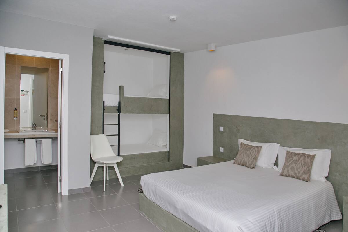 A modern bedroom with a large bed, a small white chair, a bathroom with a mirror, and a shower area.