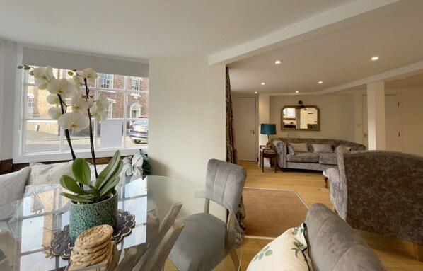 A modern living room with a glass coffee table, a grey sofa, and a dining area with a plant and a table. - Humbug Cottage (Much Wenlock)