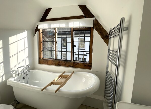 A bathroom with a freestanding bathtub, a window with a view of buildings, and a radiator. - Humbug Cottage (Much Wenlock)
