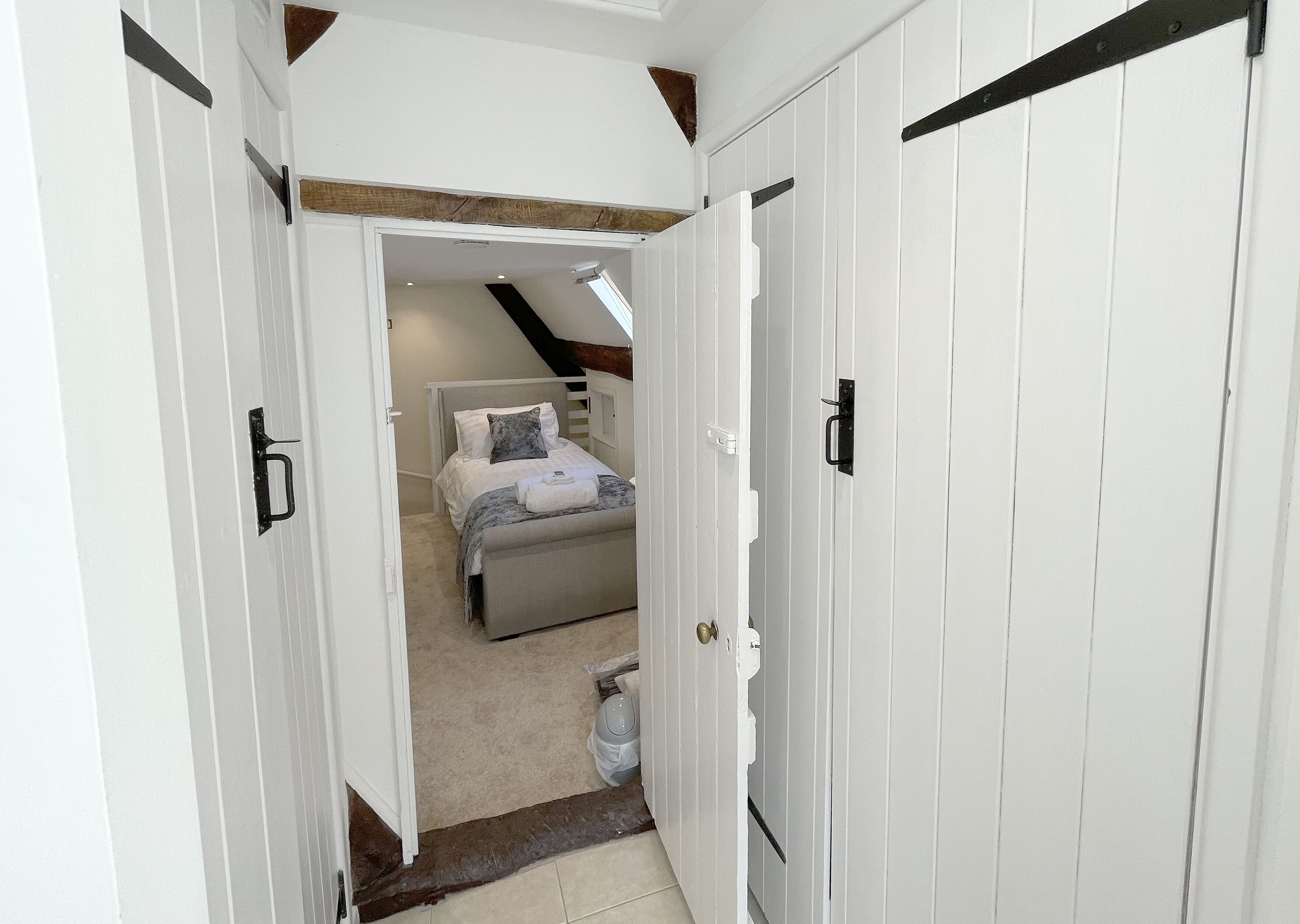 A narrow hallway leading to a bedroom with a bed, a small window, and a white door with a black handle.