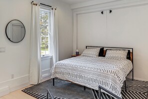 Une chambre à coucher avec un lit, une fenêtre avec des rideaux, un miroir rond et un tapis à motifs.