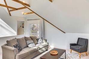 Un salon moderne avec un canapé, un fauteuil et une table basse, des poutres en bois au plafond et une fenêtre avec des rideaux.