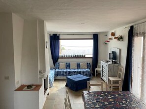 Quarto de hotel pequeno, com uma cama, escrivaninha com computador, mesa e janela com cortinas.