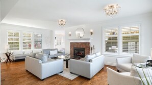 Ein geräumiges Wohnzimmer mit Kamin, weißen Sofas und einem großen Fenster mit Jalousien.