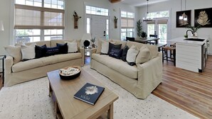 A spacious living room with two beige sofas, a coffee table, and a kitchen area with a white countertop and wooden flooring.