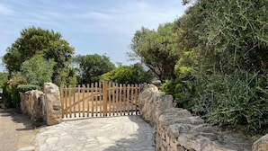 A stone pathway leading to a wooden gate surrounded by stone walls and dense greenery. - Emeraldholidaycottage Mirto - Apartment in Villa (San Pantaleo)