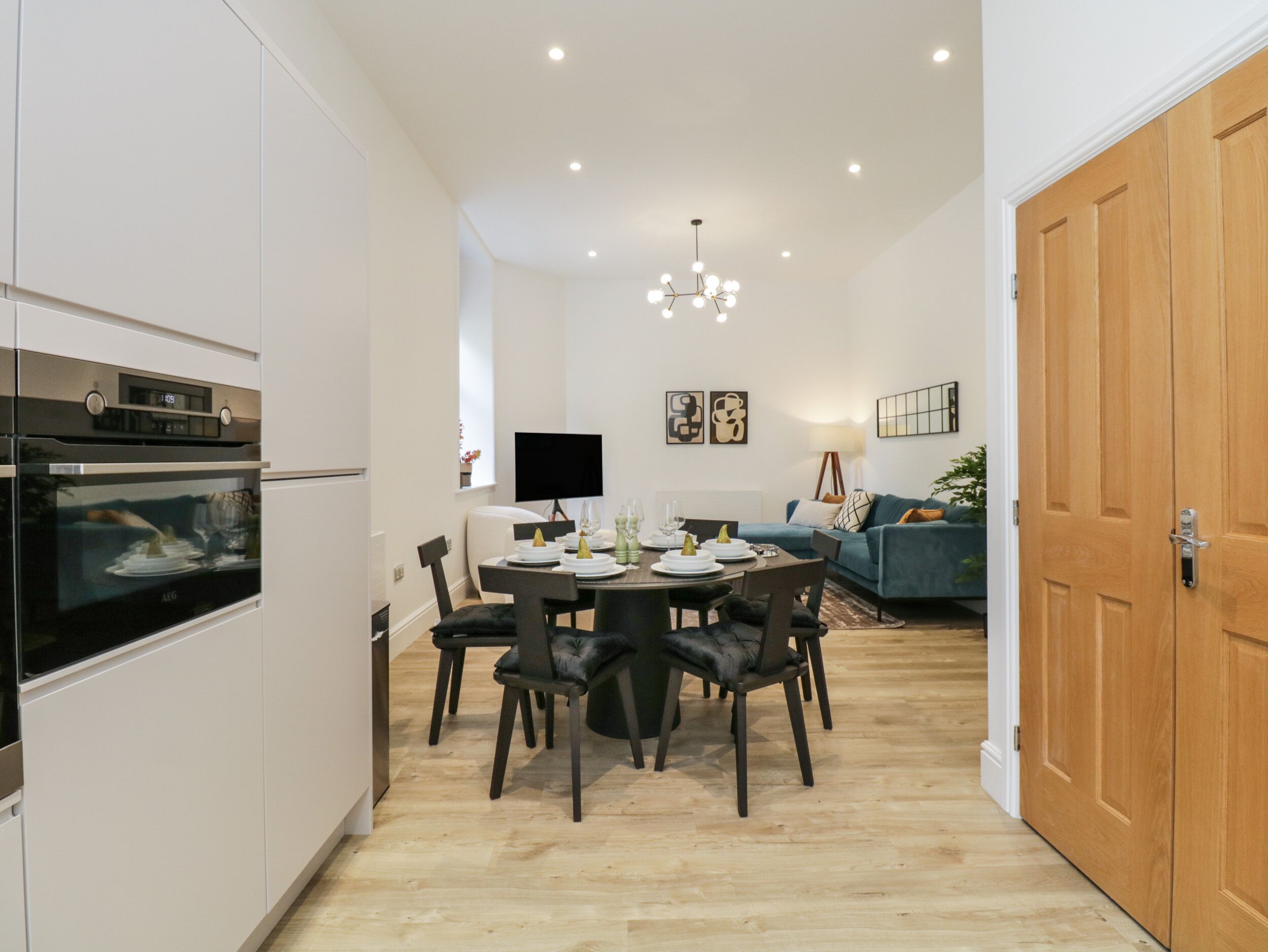 A modern kitchen with built-in appliances, a dining area with a round table and chairs, a living room with a sofa and a TV, and a wooden door.