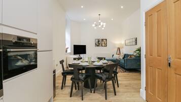 A modern kitchen with built-in appliances, a dining area with a round table and chairs, a living room with a sofa and a TV, and a wooden door.