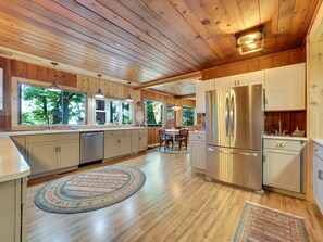 A modern kitchen with wooden cabinets, stainless steel appliances, and large windows offering views of greenery.