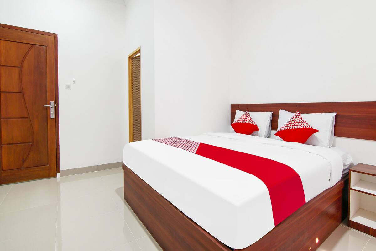A neatly made bed with white and red bedding, a wooden headboard, and a wooden nightstand with a drawer.