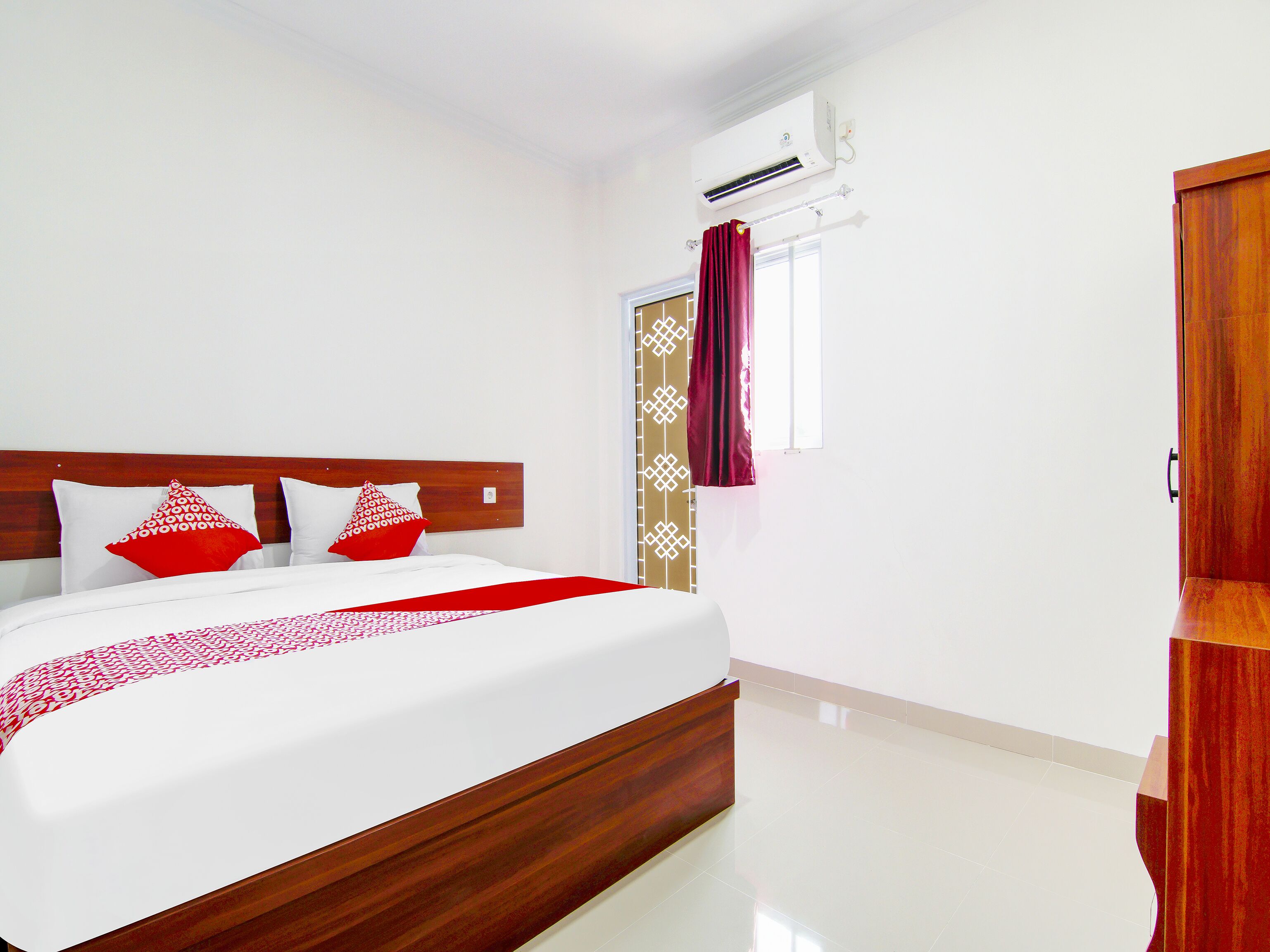 A neatly made bed with a wooden headboard, a red and white patterned throw, and a window with a curtain.