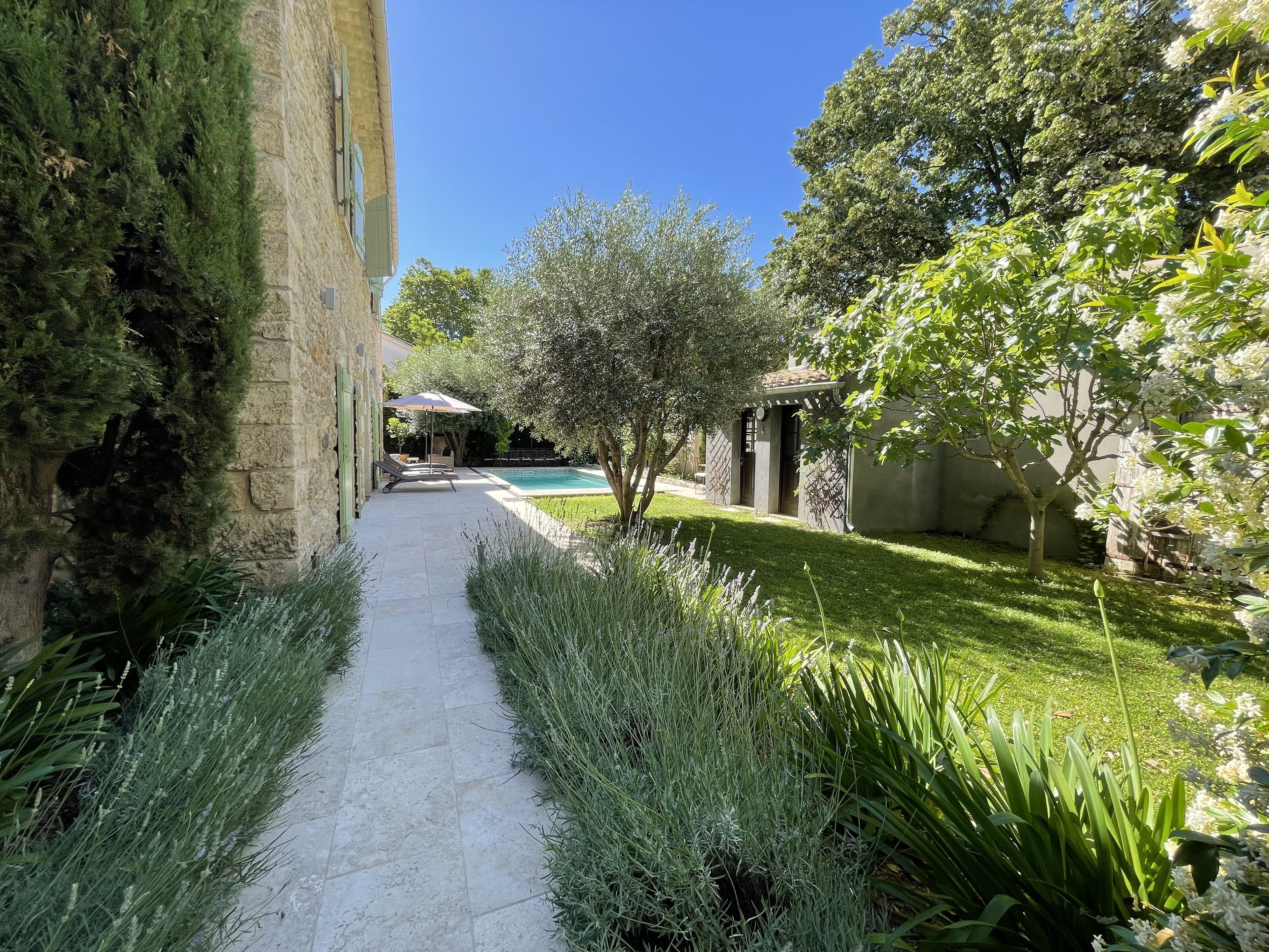 Un chemin mène à un espace piscine, avec un bâtiment d’un côté et un jardin bordé d’arbres de l’autre.