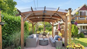 A covered patio with wicker furniture and a central table, surrounded by lush greenery and a house in the background. - LAUREL HOUSE : A Quiet Oasis in NE Portland (Portland)