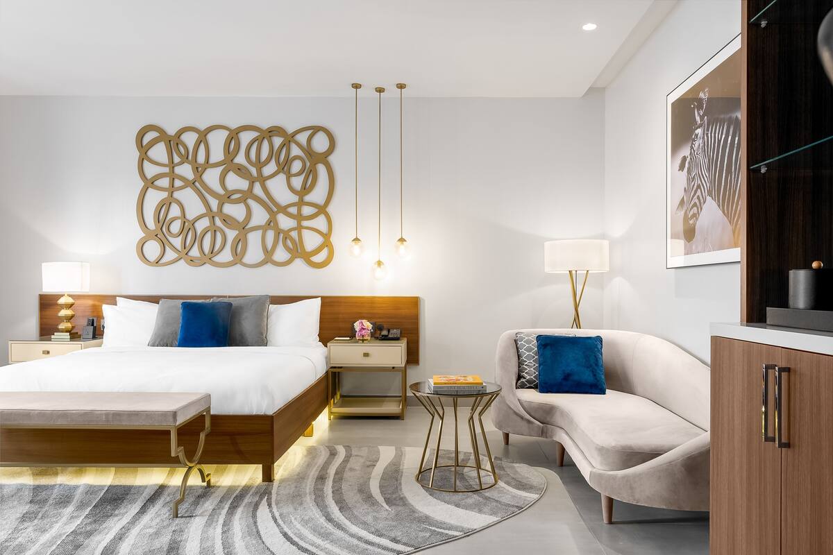 A modern hotel room with a bed, bedside table, lamp, sofa, and a decorative wall piece.