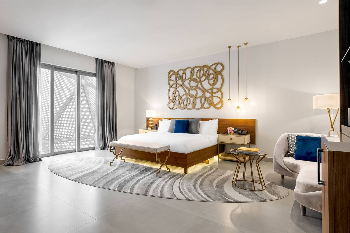 A modern hotel room with a large bed, a desk, a chair, a sofa, and a large window with curtains.