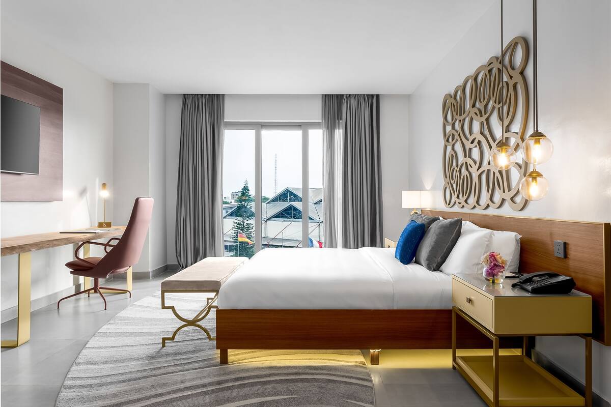 A hotel room with a bed, bedside table, lamp, chair, desk, and a large window with curtains.