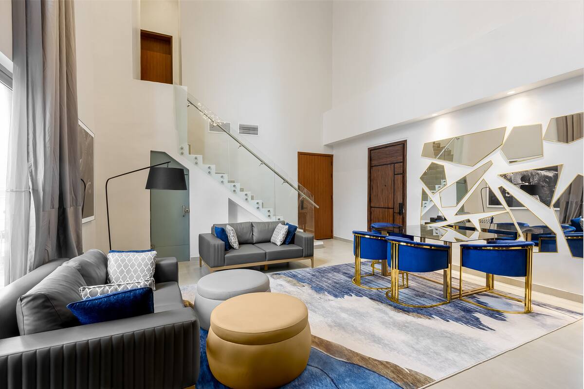 A spacious living area with a grey sofa, ottomans, and blue bar stools. A staircase with glass railings leads to an upper level.