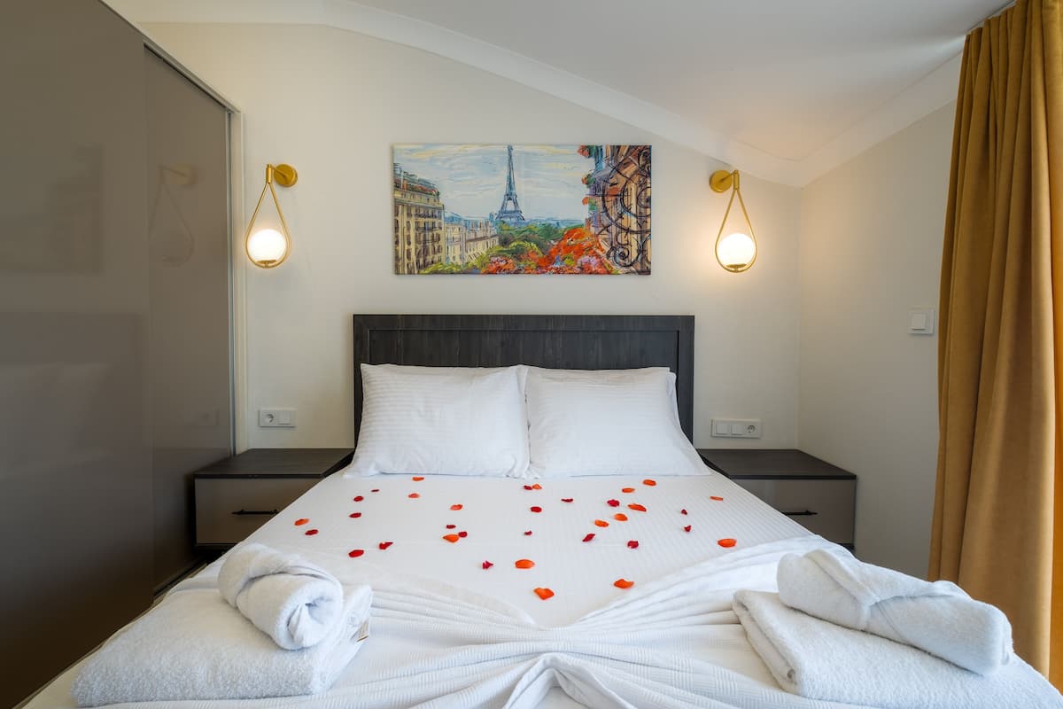 A hotel room with a bed, white linens, a painting of Paris, and two bedside lamps.