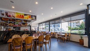 A modern restaurant interior with wooden tables and chairs, a marble counter, and large windows offering a view of the street. - Tanrise City House (Fethiye)