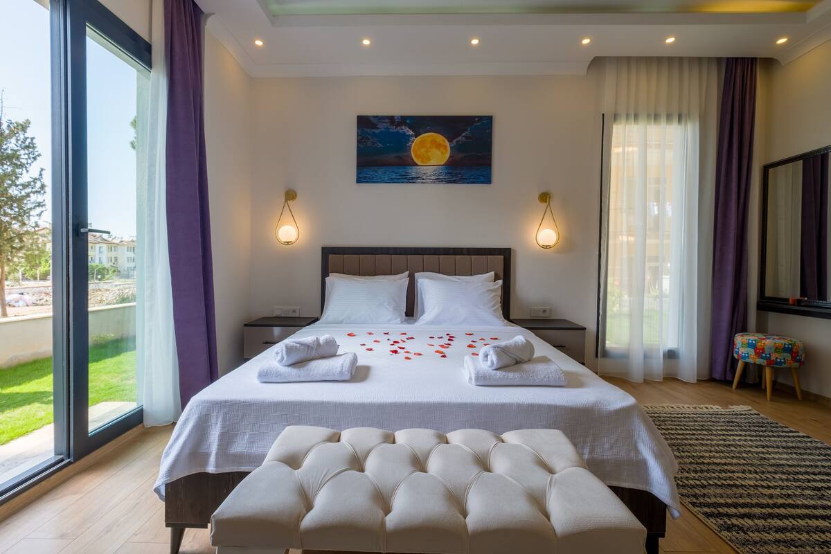 A hotel room with a large bed, a bench, a painting of a moon, and a view of the outdoors.