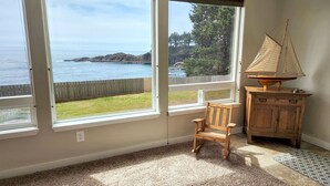 A room with large windows overlooking a coastal view, a wooden chair, and a small wooden cabinet with a sailboat model. - Ocean Front Condo! Spectacular Ocean Views!  Walk to Everything! (Depoe Bay)