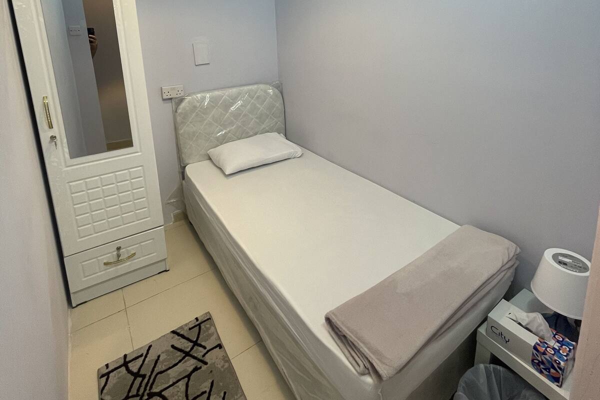 Ein kleines, ordentlich eingerichtetes Schlafzimmer mit einem Einzelbett, einem Nachttisch mit Taschentuchbox, einem Mülleimer und einem Spiegel an der Tür.
