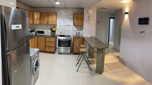 A modern kitchen with stainless steel appliances, wooden cabinets, a granite countertop, and a bar stool. - AlBarsha 1 Hostel By PHM (Dubai)