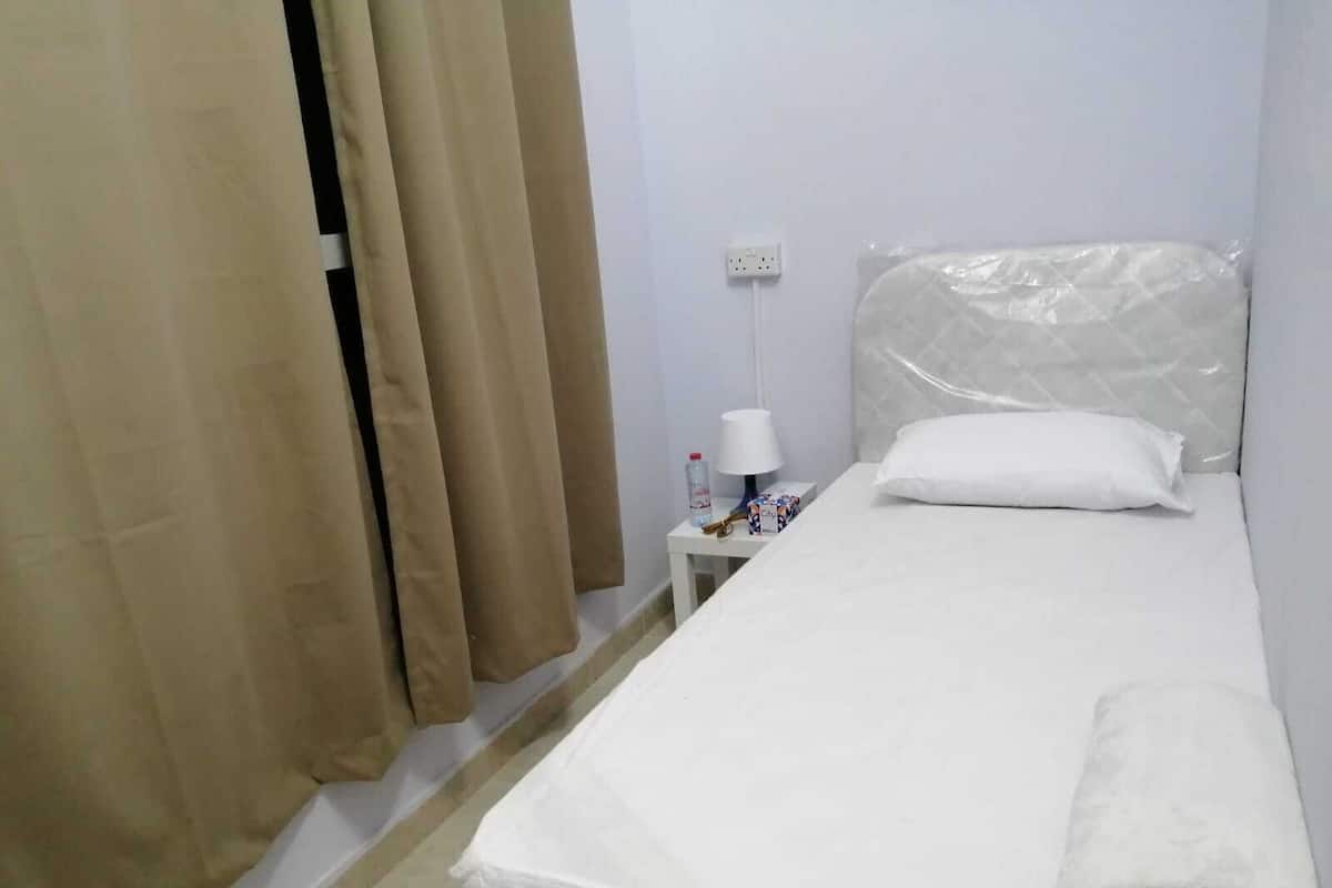 Ein Zimmer mit einem Einzelbett, einer weißen Matratze, einem weißen Kissen, einem zusammengerollten Handtuch, einem Nachttisch mit einer Lampe, einer Flasche und einer Schachtel.