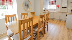 A dining area with a wooden table and chairs, a kitchen with white cabinets, and a window with plaid curtains. - North Firbank - Culzean Castle (Maybole)